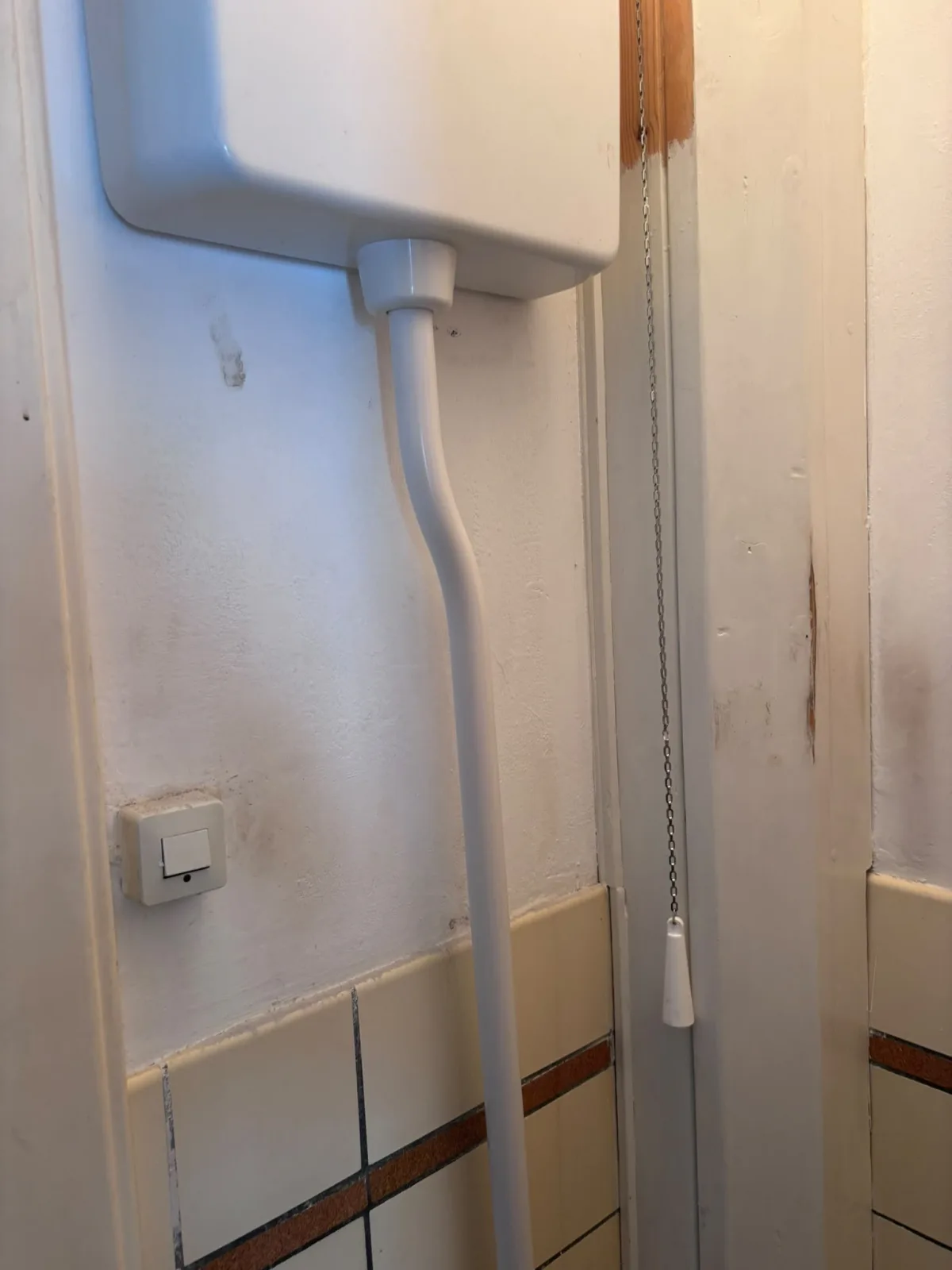 Verhoogd toiletreservoir met afvoerbuis, lichtknop en vlekken/vochtsporen op muur.