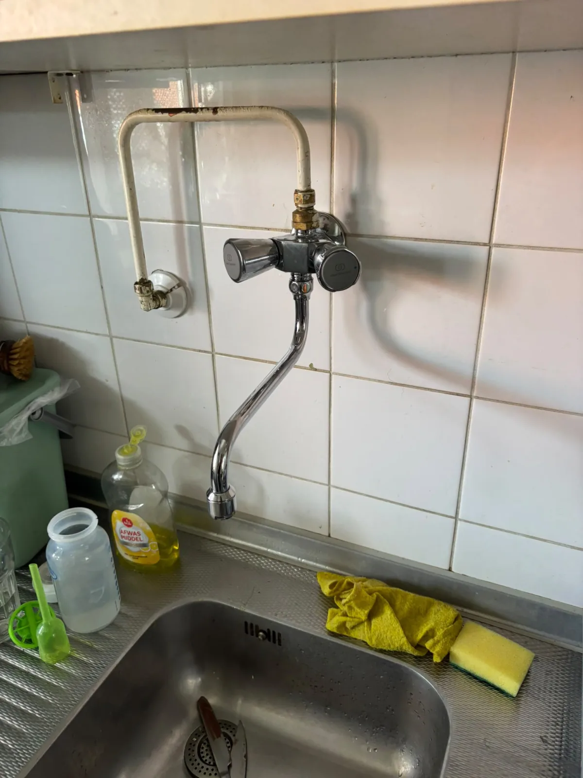 Detail gootsteen met kraan, betegelde wand en gecorrodeerde waterleidingbuis erboven.