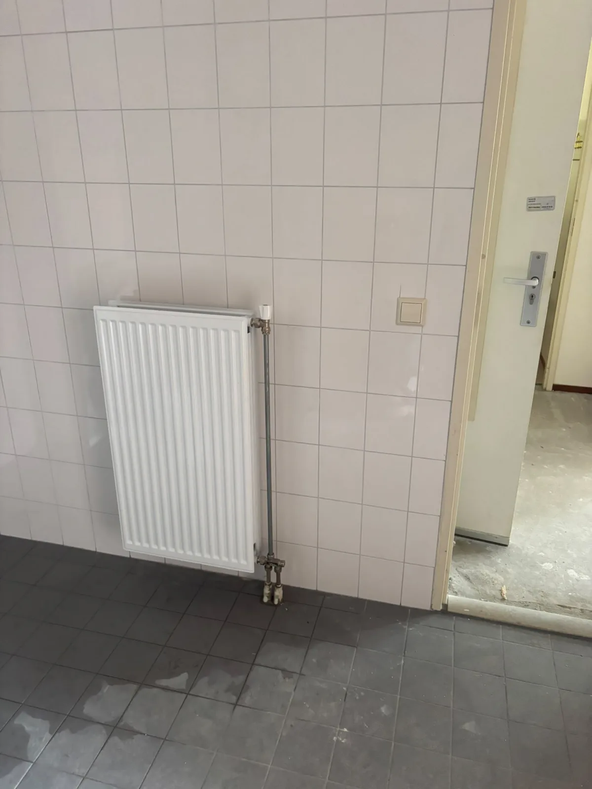 Interieurfoto met witte tegelwanden, radiator en donkergrijze tegelvloer met vochtplekken.
